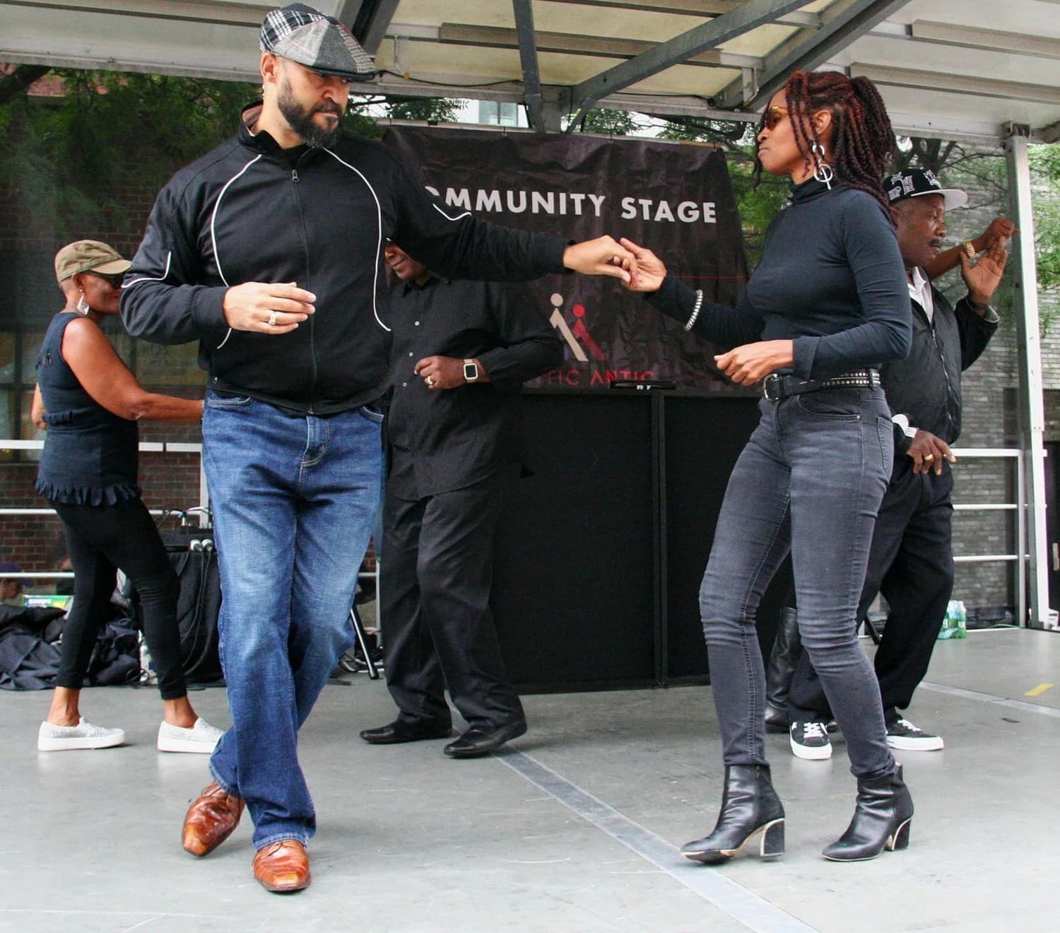 A man in a flat cap and a woman with locs stepping together mid-move on a community stage